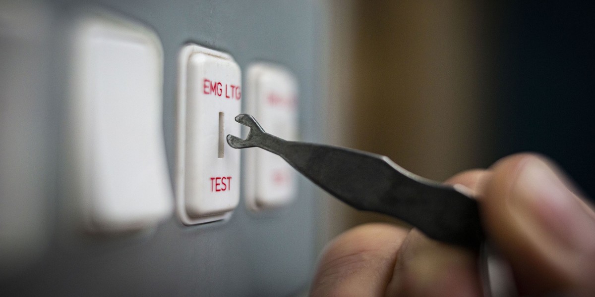 Close-up of a hand using a tool to switch an "EMG LTG TEST" button on a control panel. The scene conveys precision and focus.
